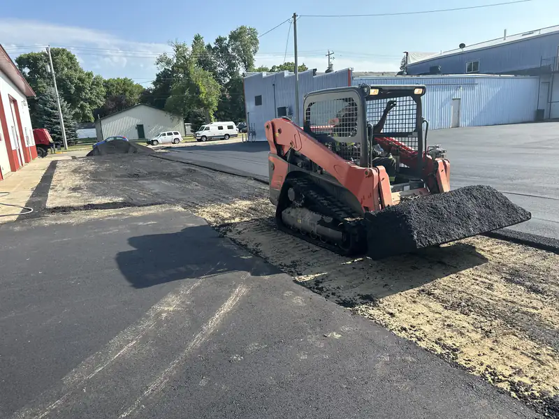 Compact track loader carrying hot asphalt mix across a prepared base on a commercial parking lot paving job