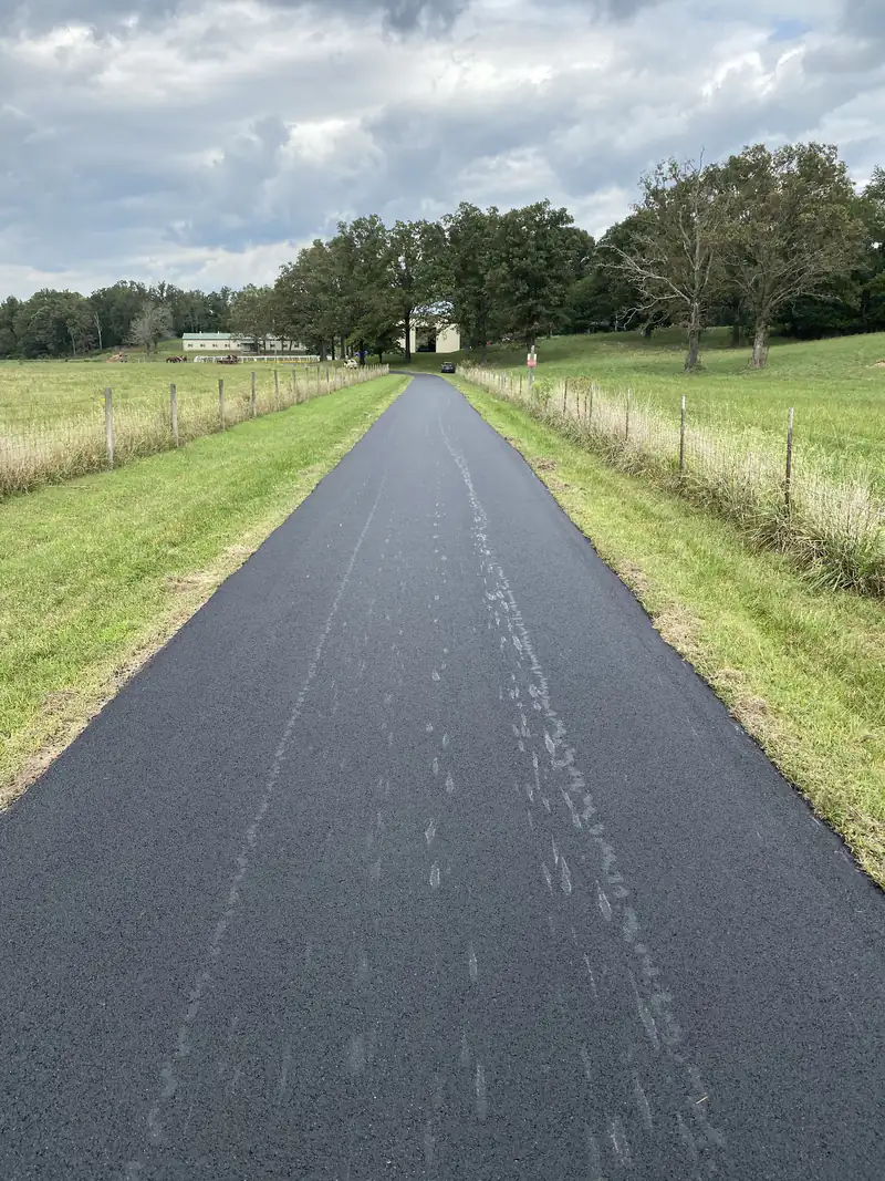 Freshly paved asphalt farm driveway running between fenced pastures toward a metal barn and mature oak trees