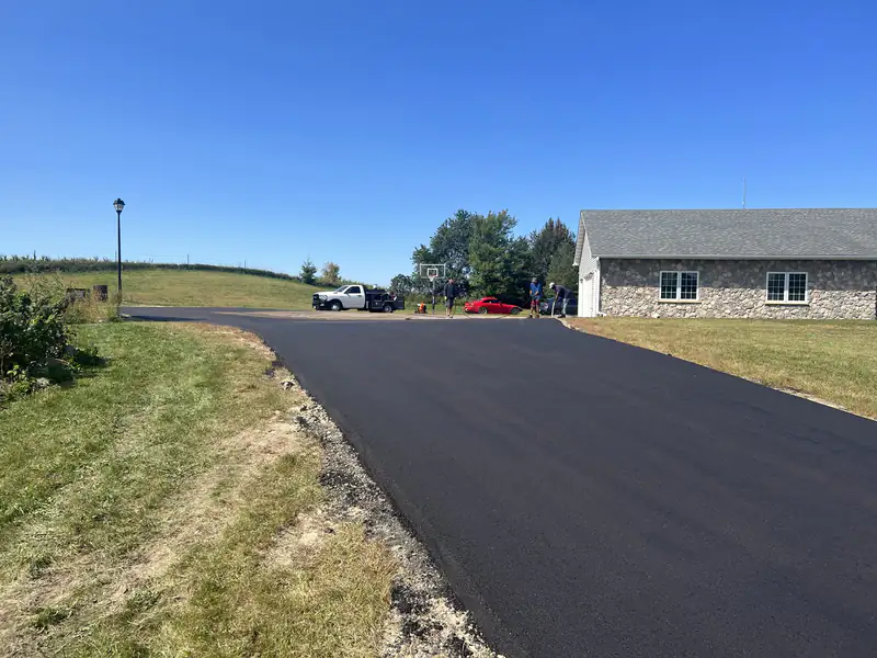 New asphalt driveway leading to a fieldstone ranch home with a cornfield and hardwood trees behind
